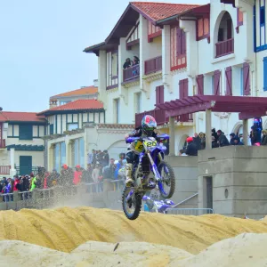 Un motorcycliste en pleine action lors de la Ronde des Sables de Hossegor, lancé à grande vitesse sur une piste de sable doré. En arrière-plan, une foule de spectateurs se tient sur la voie piétonne surélevée, observant attentivement la compétition. Le pilote, équipé d’une combinaison colorée et d’un casque intégral, soulève un nuage de sable tandis que le soleil éclaire la scène, mettant en valeur l’intensité et l’ambiance sportive de l’événement.