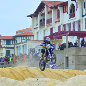 Un motorcycliste en pleine action lors de la Ronde des Sables de Hossegor, lancé à grande vitesse sur une piste de sable doré. En arrière-plan, une foule de spectateurs se tient sur la voie piétonne surélevée, observant attentivement la compétition. Le pilote, équipé d’une combinaison colorée et d’un casque intégral, soulève un nuage de sable tandis que le soleil éclaire la scène, mettant en valeur l’intensité et l’ambiance sportive de l’événement.