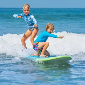 Deux enfants sur une planche de surf prenant une petite vague sur l’océan Atlantique, initiation au surf en famille dans les Landes et au Pays basque, activité nautique ludique pour découvrir les plaisirs de la glisse en toute sécurité.